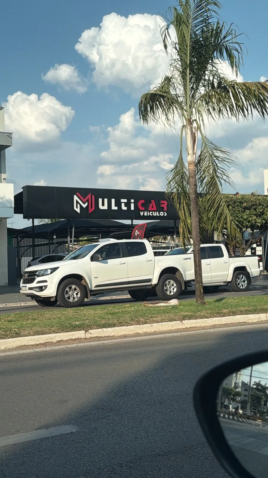 Vista da avenida: letreiro da Multicar, palmeira no canteiro e picapes no pátio